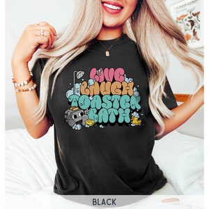 Pu&ograve; includere: Maglietta nera con un design grafico di un tostapane dei cartoni animati in una vasca da bagno con il testo "Live Laugh Toaster Bath".
