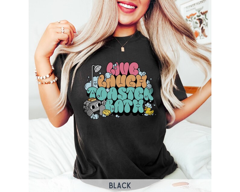 Pu&ograve; includere: Maglietta nera con un design grafico di un tostapane dei cartoni animati in una vasca da bagno con il testo "Live Laugh Toaster Bath".