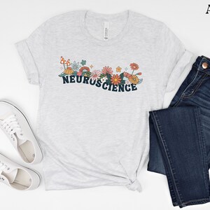May include: A light grey t-shirt with a graphic design of flowers and mushrooms. The text "NEUROSCIENCE" is printed in dark blue letters.