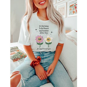 May include: White t-shirt with a graphic of two cartoon flowers with faces. The text on the shirt reads "It's Not Going To Be Flowers and Sunshine All the Time. But they will be back again."