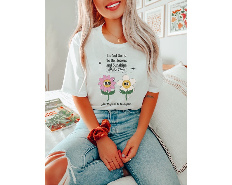 May include: White t-shirt with a graphic of two cartoon flowers with faces. The text on the shirt reads "It's Not Going To Be Flowers and Sunshine All the Time. But they will be back again."