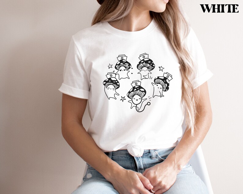 Op de afbeelding: Wit T-shirt met een afbeelding van cartoon-paddenstoelen die verpleegstersmutsen dragen. De tekst "Shroom Queens" staat op het shirt gedrukt.