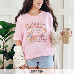 May include: A pink t-shirt with the text "It's okay if your serotonin comes in a bottle" and a cartoon illustration of a pill bottle with the text "Self Love Club" on it.