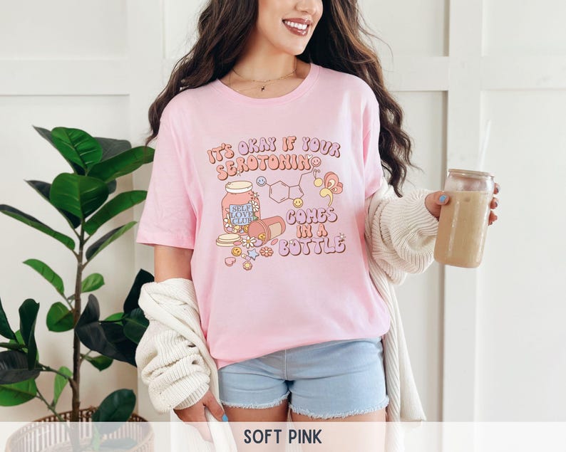 May include: A pink t-shirt with the text "It's okay if your serotonin comes in a bottle" and a cartoon illustration of a pill bottle with the text "Self Love Club" on it.