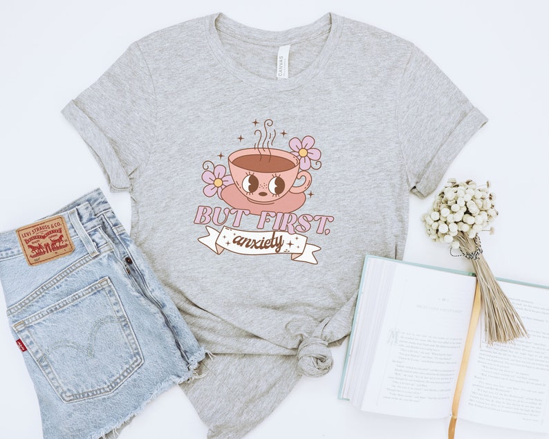 May include: Gray t-shirt with a pink and white cartoon coffee cup with the text "BUT FIRST, anxiety" on a white banner.