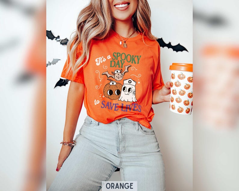 Puede incluir: Camiseta naranja con el texto "It's a SPOOKY DAY to SAVE LIVES" y gr&aacute;ficos tem&aacute;ticos de Halloween. El dise&ntilde;o incluye un murci&eacute;lago, una calabaza y un fantasma. Tambi&eacute;n se ve una taza de caf&eacute; blanca y naranja con gr&aacute;ficos de calabazas.