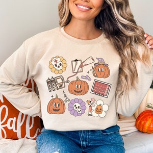 May include: A beige sweatshirt with a Halloween-themed design featuring pumpkins, a skull, a stethoscope, a heart monitor, and other medical symbols.