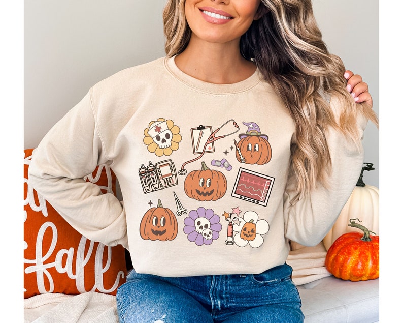 May include: A beige sweatshirt with a Halloween-themed design featuring pumpkins, a skull, a stethoscope, a heart monitor, and other medical symbols.