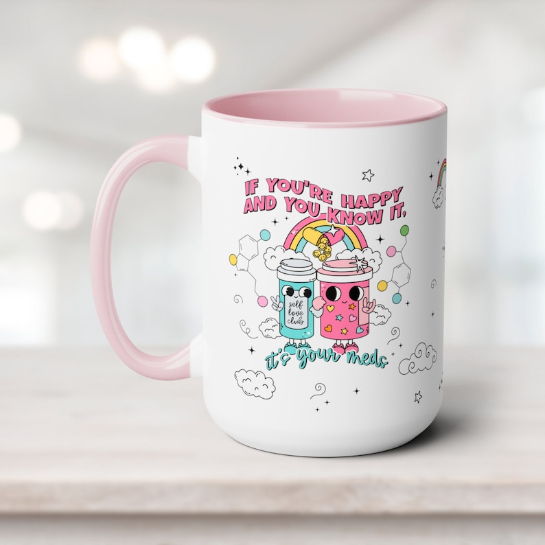 May include: White ceramic mug with a pink rim and handle. The mug features a colourful illustration of two pill bottles with faces and the text "If you're happy and you know it, it's your meds."