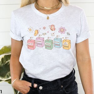 May include: A light grey t-shirt with a graphic of six colorful juice boxes. Each juice box has a different positive affirmation printed on it, including "Self Love Juice", "Dopamine Juice", "Empathy Juice", "Gratitude Juice", and "Anti Anxiety Juice".