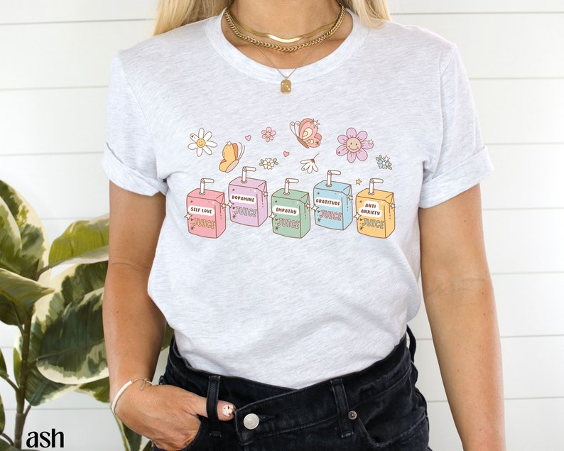 May include: A light grey t-shirt with a graphic of six colorful juice boxes. Each juice box has a different positive affirmation printed on it, including "Self Love Juice", "Dopamine Juice", "Empathy Juice", "Gratitude Juice", and "Anti Anxiety Juice".