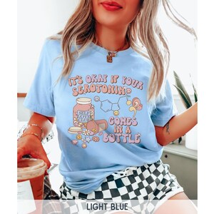 May include: Light blue t-shirt with a graphic of a pill bottle with the text "It's okay if your serotonin comes in a bottle" and "Self Love Club" on the label.