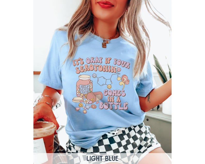 May include: Light blue t-shirt with a graphic of a pill bottle with the text "It's okay if your serotonin comes in a bottle" and "Self Love Club" on the label.