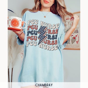 K&ouml;nnte beinhalten: Hellblaues T-Shirt mit einem Grafikdruck eines rot-wei&szlig;-blauen Blitzes und dem Text "PCU NURSE", der in einer Retro-Schrift wiederholt wird.