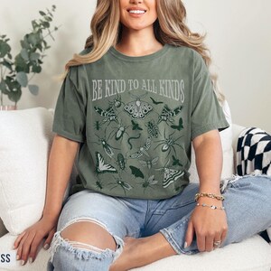Op de afbeelding: Mosgroen t-shirt met de tekst "BE KIND TO ALL KINDS" en insectenillustraties. Het ontwerp toont verschillende insecten, waaronder vlinders, motten en spinnen. Het shirt is een comfortabele, casual stijl.