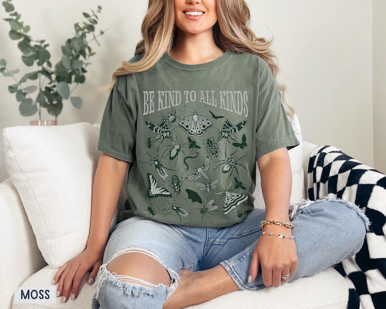 Op de afbeelding: Mosgroen t-shirt met de tekst "BE KIND TO ALL KINDS" en insectenillustraties. Het ontwerp toont verschillende insecten, waaronder vlinders, motten en spinnen. Het shirt is een comfortabele, casual stijl.