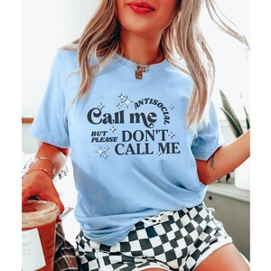 Puede incluir: Una camiseta azul claro con un gr&aacute;fico negro que dice "Call me antisocial but please don't call me". El gr&aacute;fico est&aacute; rodeado de peque&ntilde;as estrellas.