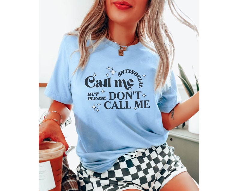Puede incluir: Una camiseta azul claro con un gr&aacute;fico negro que dice "Call me antisocial but please don't call me". El gr&aacute;fico est&aacute; rodeado de peque&ntilde;as estrellas.