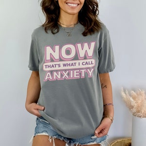 May include: A gray t-shirt with the text "NOW THAT'S WHAT I CALL ANXIETY" in pink and white lettering. The shirt is a casual style, suitable for everyday wear. The overall aesthetic is relaxed and comfortable.