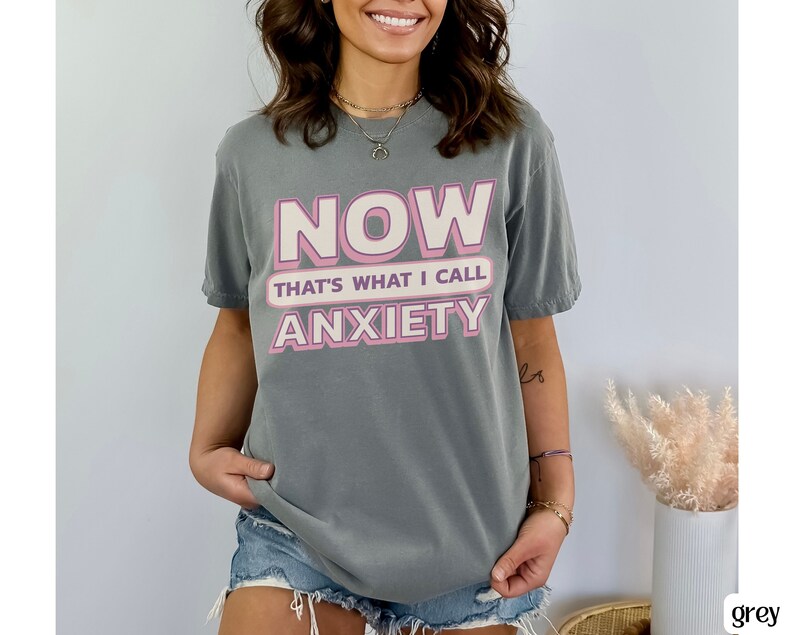 May include: A gray t-shirt with the text "NOW THAT'S WHAT I CALL ANXIETY" in pink and white lettering. The shirt is a casual style, suitable for everyday wear. The overall aesthetic is relaxed and comfortable.