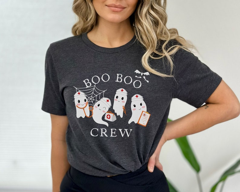 May include: A dark grey t-shirt featuring a white graphic of three ghosts dressed as nurses. The ghosts are holding medical supplies and the text "BOO BOO CREW" is printed below them.