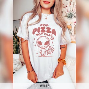 May include: White t-shirt with a graphic of an alien holding a slice of pizza. The text "Pop Pizza Not Pills" is printed on the shirt.