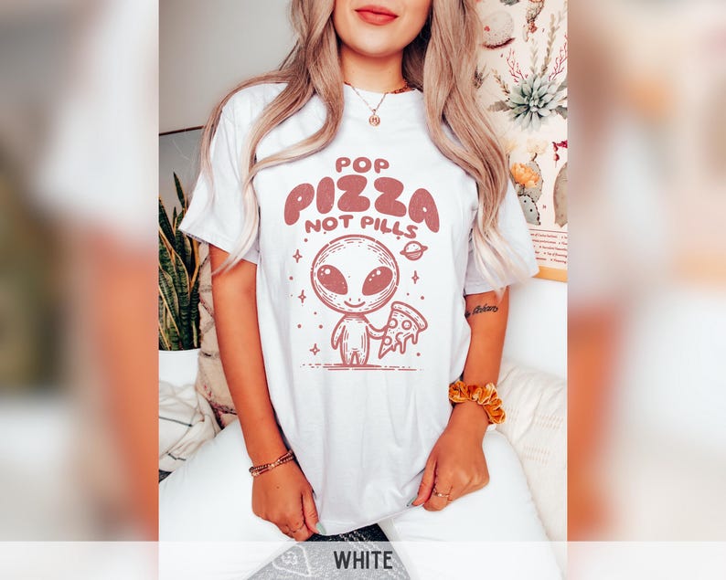 May include: White t-shirt with a graphic of an alien holding a slice of pizza. The text "Pop Pizza Not Pills" is printed on the shirt.