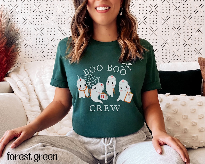 May include: A forest green t-shirt featuring a design of three ghosts dressed as nurses. The ghosts are wearing red nurse hats and holding medical equipment. The text "BOO BOO CREW" is printed on the shirt.