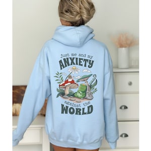 May include: Light blue hoodie with a graphic of a frog and snail on mushrooms. The text on the hoodie reads "Just me and my anxiety against the world."