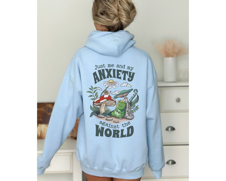 May include: Light blue hoodie with a graphic of a frog and snail on mushrooms. The text on the hoodie reads "Just me and my anxiety against the world."