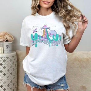 May include: White t-shirt with a retro space-themed graphic. The design features a UFO, rocket, cactus, and the text "LET'S GET LOST". The shirt has a relaxed fit and is made of a soft material.