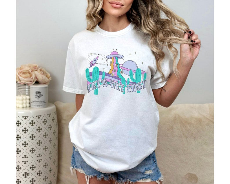 May include: White t-shirt with a retro space-themed graphic. The design features a UFO, rocket, cactus, and the text "LET'S GET LOST". The shirt has a relaxed fit and is made of a soft material.