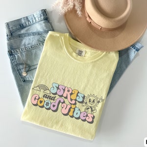May include: A light yellow t-shirt with the text "SSRS and Good Vibes" in a retro font, featuring a rainbow, sun, and flower graphic. The shirt is paired with light blue jeans and a tan hat.