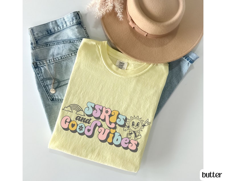 May include: A light yellow t-shirt with the text "SSRS and Good Vibes" in a retro font, featuring a rainbow, sun, and flower graphic. The shirt is paired with light blue jeans and a tan hat.