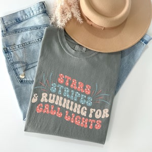 May include: A grey t-shirt with the text "Stars Stripes & Running for Call Lights" in a retro font with red, blue, and white accents. The shirt is folded and laid on a pair of blue jeans and a brown hat.