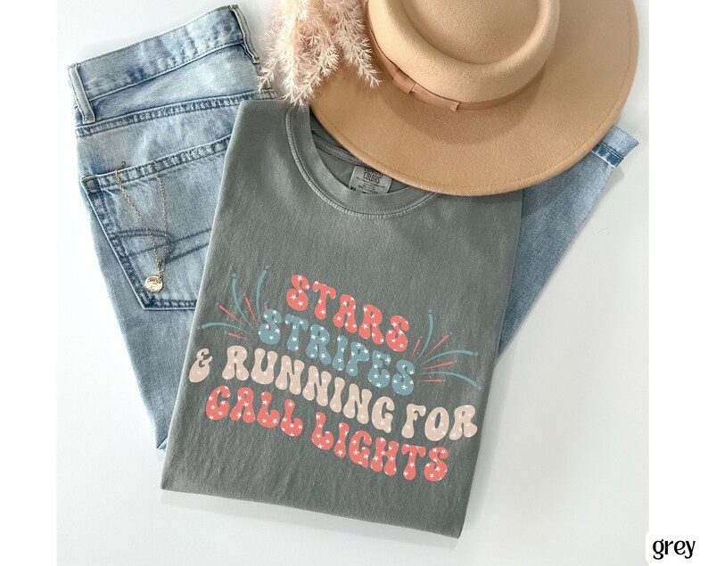 May include: A grey t-shirt with the text "Stars Stripes & Running for Call Lights" in a retro font with red, blue, and white accents. The shirt is folded and laid on a pair of blue jeans and a brown hat.