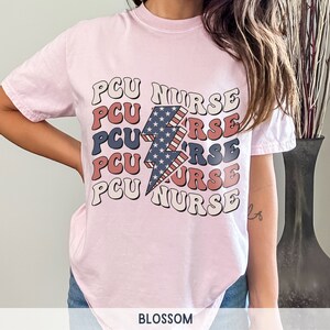 K&ouml;nnte beinhalten: Rosa T-Shirt mit einem Grafikdruck eines Blitzes mit einem amerikanischen Flaggenmuster und dem Text "PCU NURSE", der f&uuml;nfmal wiederholt wird.