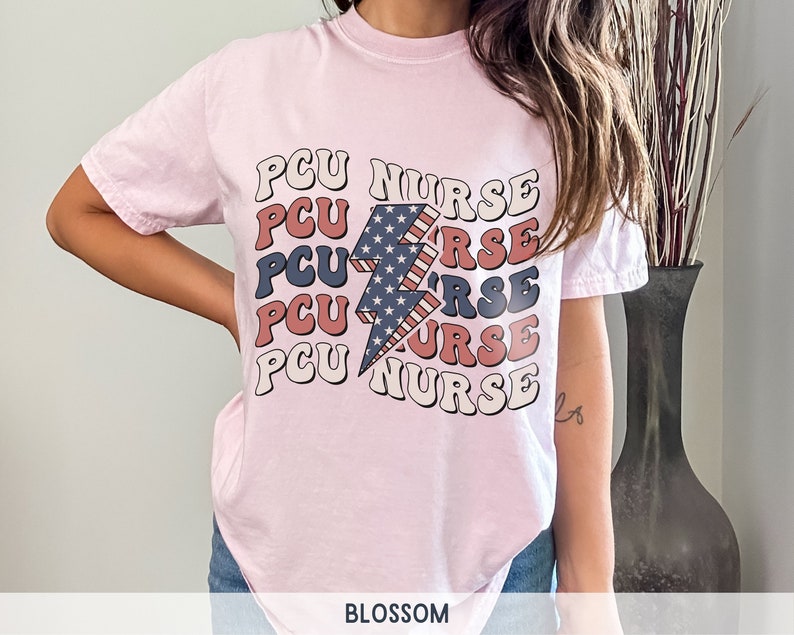 K&ouml;nnte beinhalten: Rosa T-Shirt mit einem Grafikdruck eines Blitzes mit einem amerikanischen Flaggenmuster und dem Text "PCU NURSE", der f&uuml;nfmal wiederholt wird.