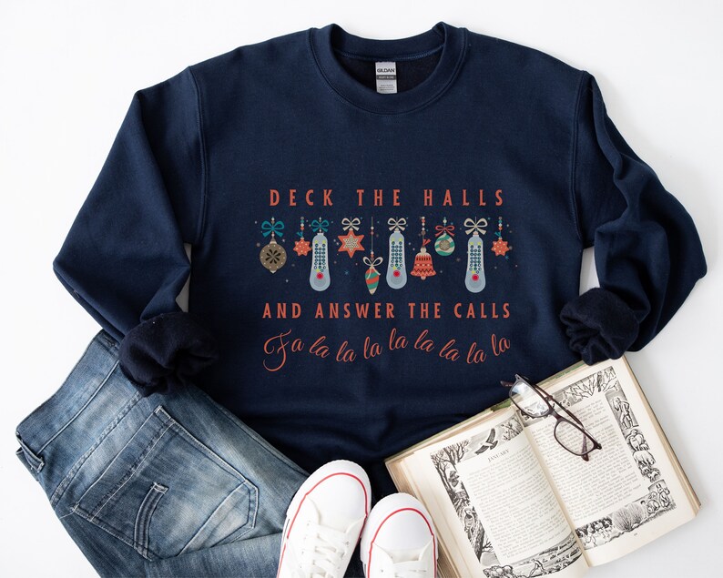 Może przedstawiać: Granatowa bluza z napisem "Deck the Halls and Answer the Calls" w kolorze czerwonym. Tekst jest otoczony rzędem kolorowych ozd&oacute;b choinkowych.