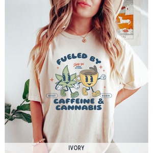 Puede incluir: Una camiseta de color crema con un gr&aacute;fico de una taza de caf&eacute; de dibujos animados y una hoja de marihuana. El texto de la camiseta dice "Fueled by Caffeine & Cannabis. They go hand in hand. Best Buds."