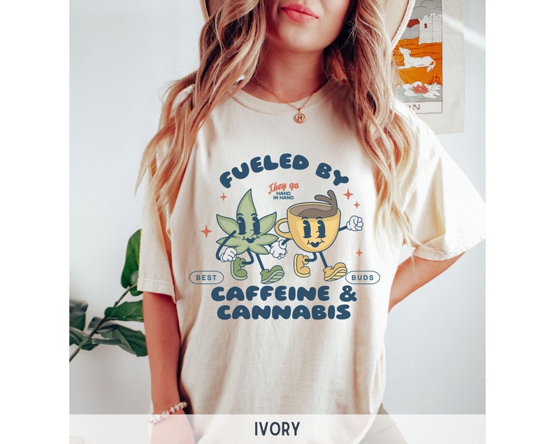 Puede incluir: Una camiseta de color crema con un gr&aacute;fico de una taza de caf&eacute; de dibujos animados y una hoja de marihuana. El texto de la camiseta dice "Fueled by Caffeine & Cannabis. They go hand in hand. Best Buds."