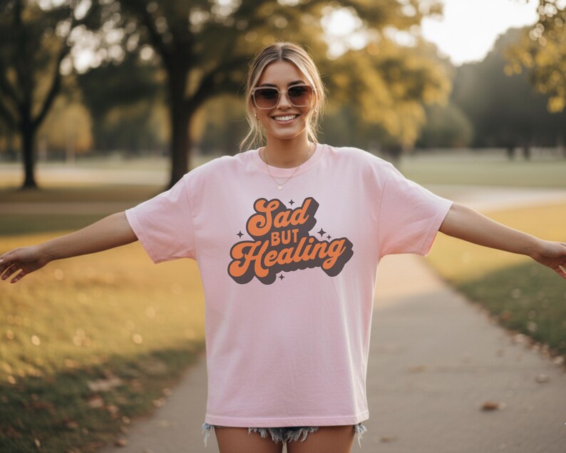 Puede incluir: Una camiseta rosa claro con las palabras "Sad but Healing" en un dise&ntilde;o retro naranja y marr&oacute;n. La camiseta es de corte holgado y tiene mangas cortas. La persona lleva gafas de sol y pantalones cortos.