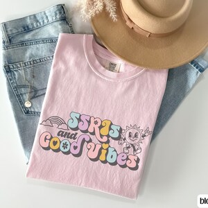 May include: A light pink t-shirt with the text "SSRS and Good Vibes" in a retro font, featuring a rainbow and a smiling sun graphic. The shirt is paired with light wash jeans and a tan hat.