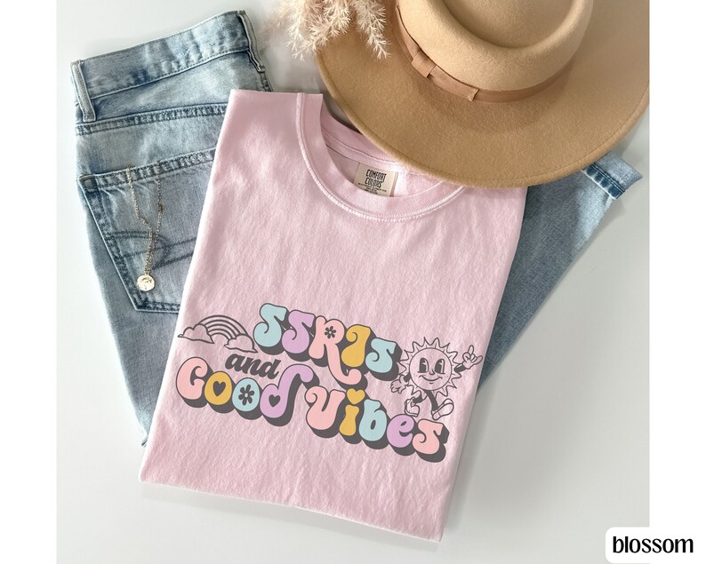 May include: A light pink t-shirt with the text "SSRS and Good Vibes" in a retro font, featuring a rainbow and a smiling sun graphic. The shirt is paired with light wash jeans and a tan hat.