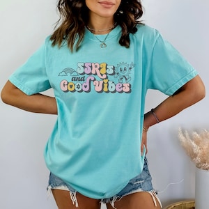May include: A light blue t-shirt with the text "SSRIs and Good Vibes" in a retro font. The graphic includes a rainbow, a smiling sun, and a flower. The shirt is a casual, oversized style.