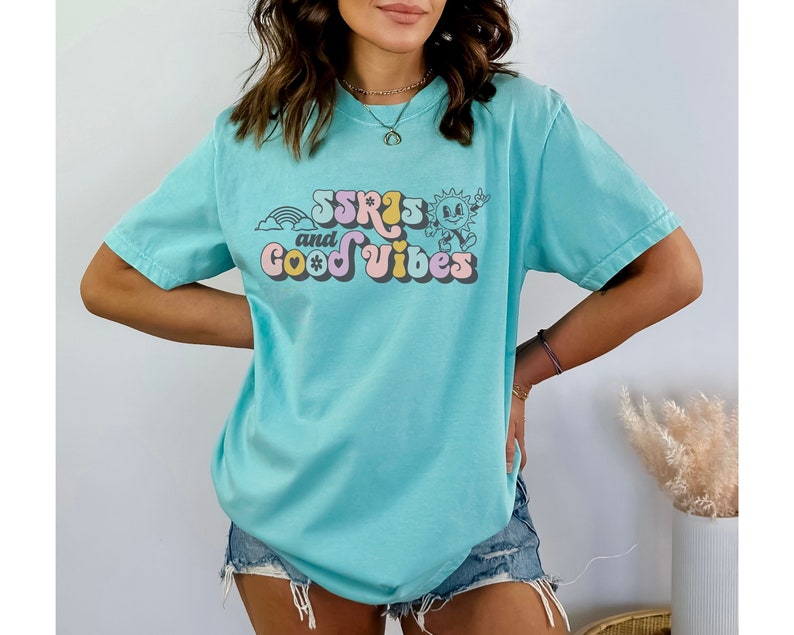May include: A light blue t-shirt with the text "SSRIs and Good Vibes" in a retro font. The graphic includes a rainbow, a smiling sun, and a flower. The shirt is a casual, oversized style.