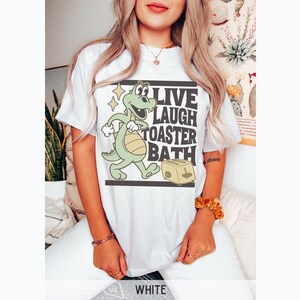 May include: White t-shirt featuring a cartoon dinosaur with the text "LIVE LAUGH TOASTER BATH". The dinosaur is green and yellow, and the toaster is beige. The shirt is a casual, unisex style.