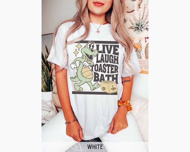 May include: White t-shirt featuring a cartoon dinosaur with the text "LIVE LAUGH TOASTER BATH". The dinosaur is green and yellow, and the toaster is beige. The shirt is a casual, unisex style.