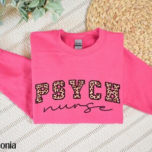 May include: A bright pink sweatshirt with the words "PSYCH nurse" printed on the front. The word "PSYCH" is in a bold, block letter style with a leopard print pattern. The word "nurse" is in a cursive font.