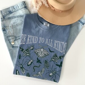 Op de afbeelding: Een blauw T-shirt met de tekst "BE KIND TO ALL KINDS" en insectenillustraties. Het shirt wordt gecombineerd met een spijkerbroek en een beige hoed. De afbeelding heeft een casual, ontspannen uitstraling.